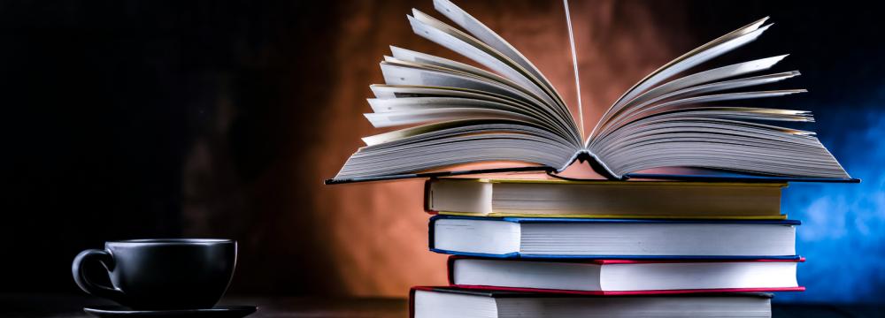 Une pile de livres, dont l'un est ouvert, les pages étalées en éventail, à côté d'une tasse de café posée sur une table.