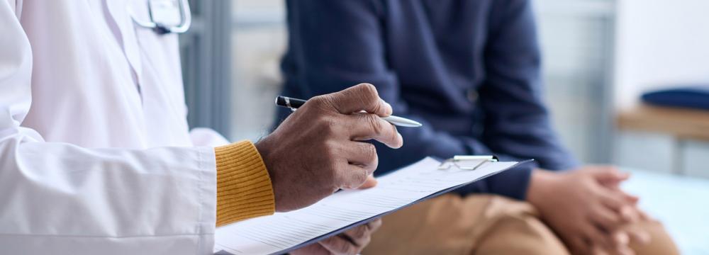 A doctor in a white lab coat with a stethoscope around his neck is holding a writing pad and a pen with a patient dressed in a long-sleeved dark blue shirt and brown pants seated in a clinic room. 