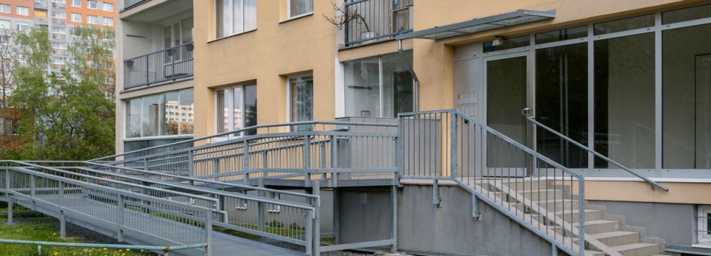 An exterior brown apartment building with an accessibility ramp, alongside stairs, and grey handrails leading to the entrance.