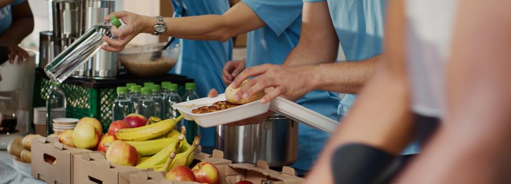 Behind a long table, a handful of volunteers in t-shirts with the word "Volunteer" printed on them, distribute fresh fruits that include apples and bananas, and green legumes.