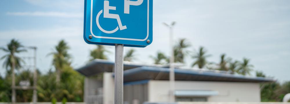 Panneau de stationnement accessible bleu avec un symbole de fauteuil roulant dans un parc de stationnement, avec des voitures garées, des palmiers et un bâtiment bas en arrière-plan.