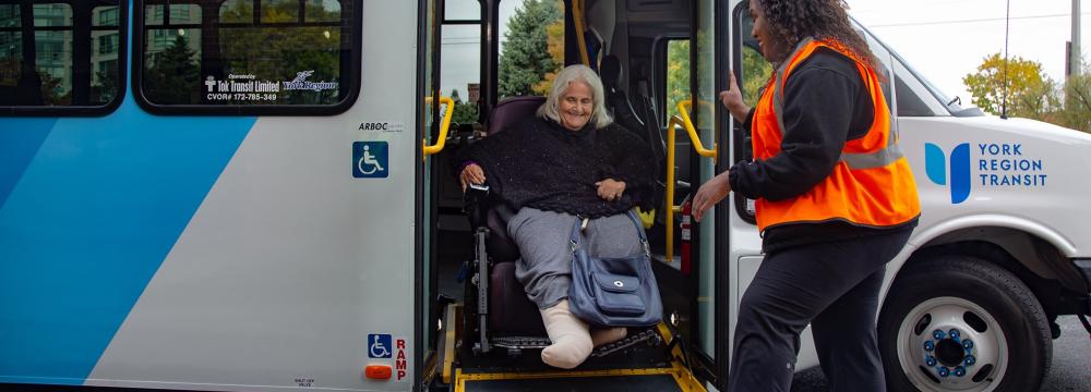 A public bus with an accessibility ramp extended as a person in a wheelchair gets off with the assistance of a transit worker.