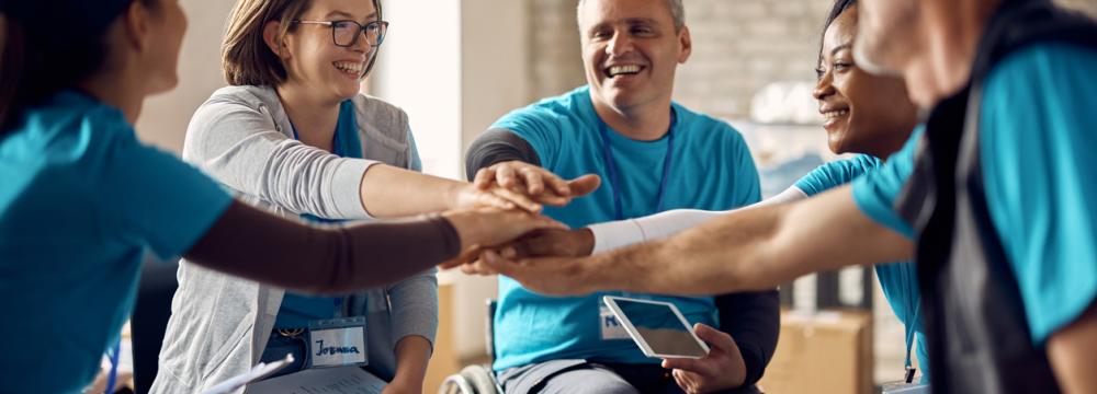 A multiracial team in a circle, including a person in a wheelcairr, gathering hands in the centre, symbolizing collaboration and unity.