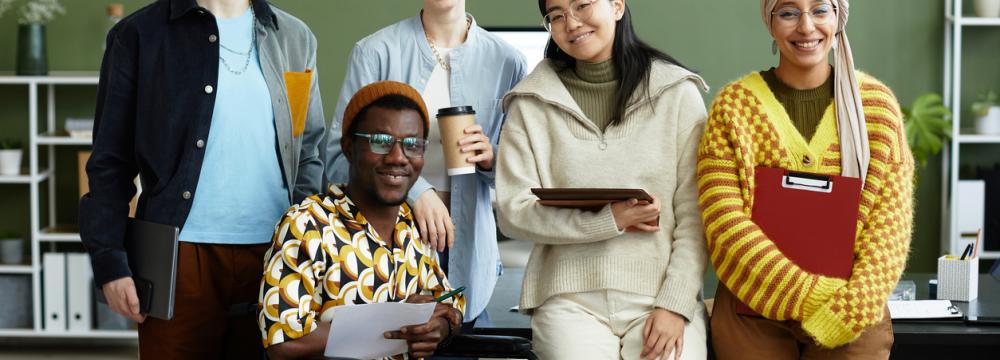 A multicultural group of employees in front of a black desk, holding papers and clipboards, four of them standing and one in a wheelchair.