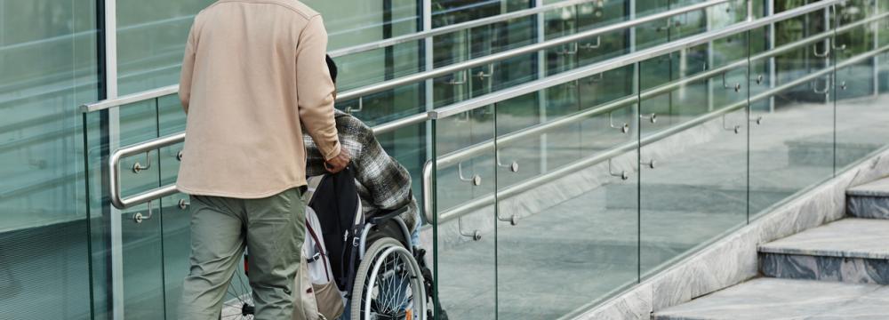 A man pushing someone in a wheelchair along a ramp toward the entrance of a glass building.