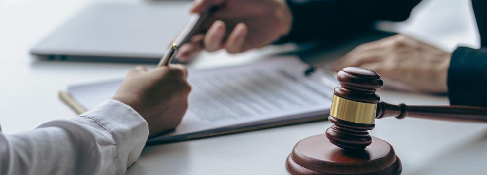 Deux personnes sont assises de chaque côté d’un bureau, un stylo à la main, prêtes à signer un document posé entre elles, à côté d’un marteau du juge.