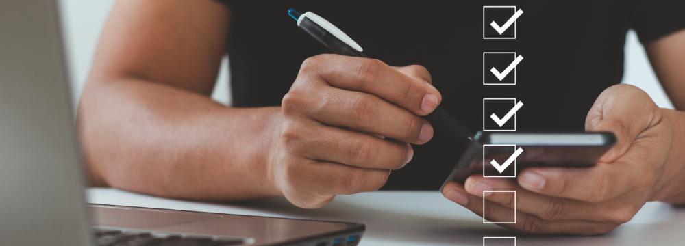 Une personne munie d'un stylo vérifie les éléments d'une liste sur un téléphone portable, tandis qu'un ordinateur portable est posé sur le bureau et des icônes de vérification numérique apparaissent superposées.