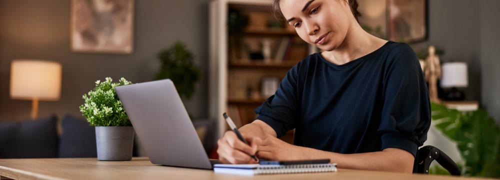 Caucasian woman in a wheelchair, working at a table, writing with a pen in a notebook, with a laptop open in front of her, in a modern home environment (with a floor lamp and a bookcase in the background).