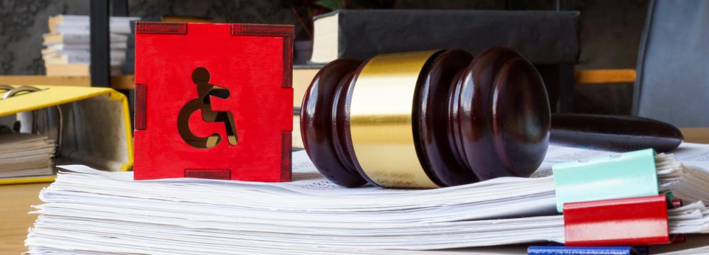 A red accessibility symbol and a gavel are placed next to each other on top of clipped papers on a brown desk.