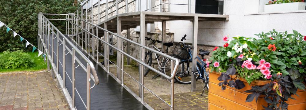 A grey accessible ramp with white metal railings leading up to the entrance of a white building, next to a flower planter