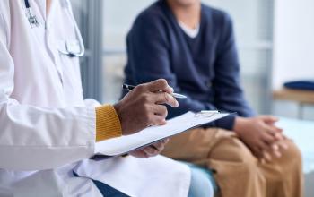 A doctor in a white lab coat with a stethoscope around his neck is holding a writing pad and a pen with a patient dressed in a long-sleeved dark blue shirt and brown pants seated in a clinic room. 