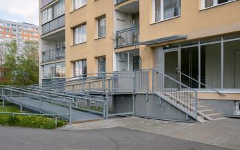 An exterior brown apartment building with an accessibility ramp, alongside stairs, and grey handrails leading to the entrance.
