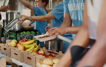 Behind a long table, a handful of volunteers in t-shirts with the word "Volunteer" printed on them, distribute fresh fruits that include apples and bananas, and green legumes.