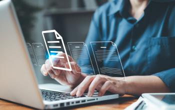 A person dressed in a blue shirt with buttons is typing on a laptop with digital graphics of documents and folders overlaid.