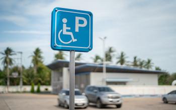 A blue accessible parking sign with a wheelchair symbol in a parking lot, with parked cars, palm trees, and a low building in the background.