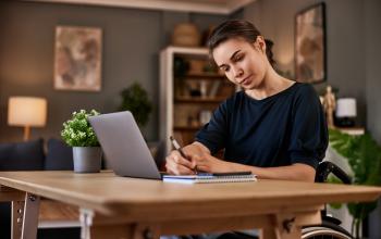 Caucasian woman in a wheelchair, working at a table, writing with a pen in a notebook, with a laptop open in front of her, in a modern home environment (with a floor lamp and a bookcase in the background).